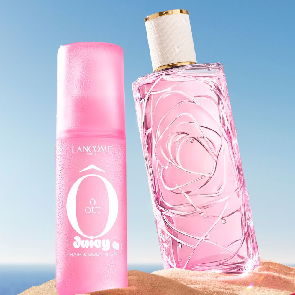 A light pink perfume bottle with a textured glass design, against a light blue sky background, with water droplets splashing around it. The bottle is seen through a circular opening in a textured pink wall, and there are blurred pink flowers in the foreground and background.